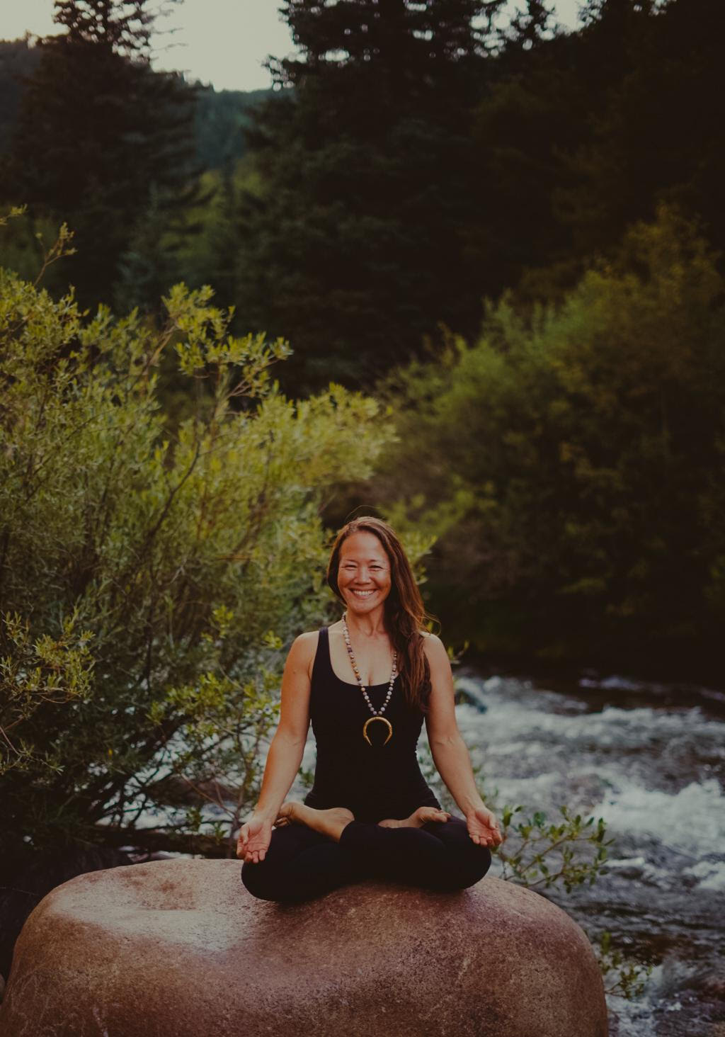 Mary Kirkendoll seated in lotus pose on a rock by a river